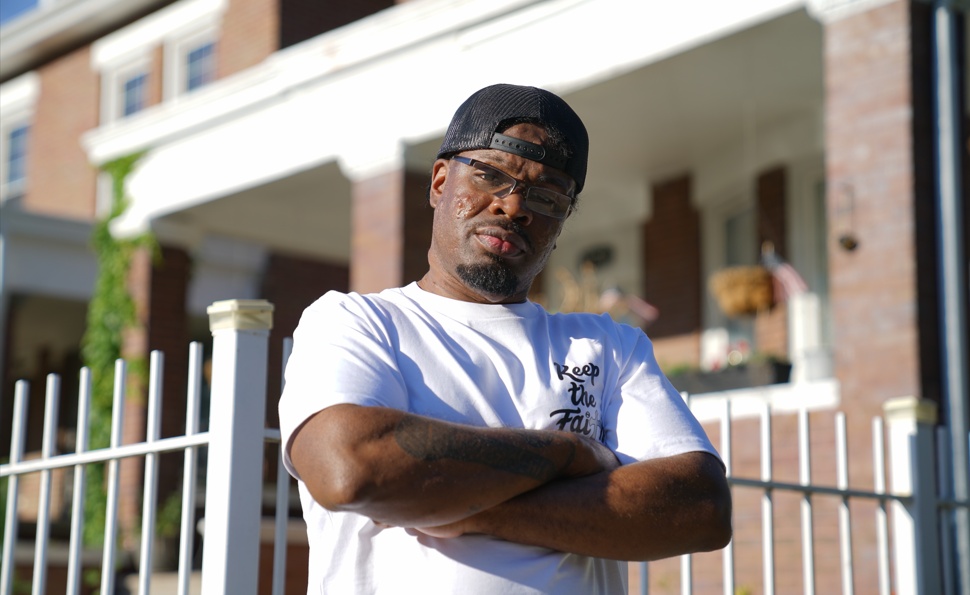 Cloak The Scribe wearing glasses, a backward black cap and a white shirt, with his arms folded outdoors.