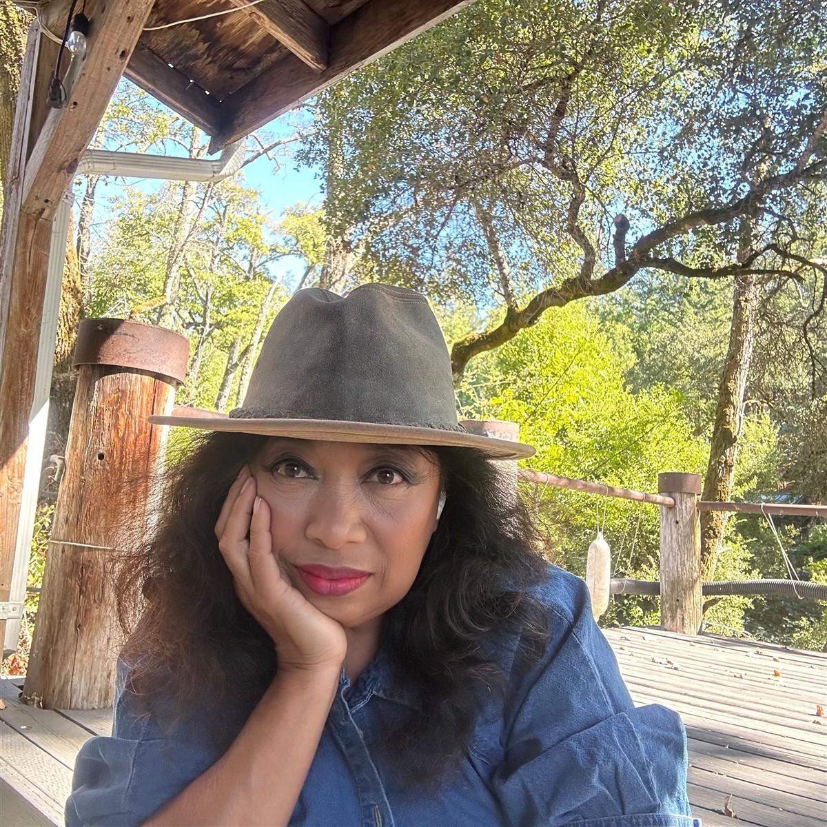 Michelle Bond, known as Michellar, wearing a wide-brimmed hat and denim shirt on a wooden deck among trees.